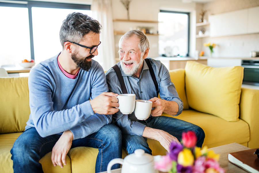 An adult son and senior father sitting on sofa indoors at home, drinking tea.