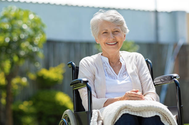 Happy senior woman sitting on wheelchair and recovering from illness. Handicapped mature woman sitting in wheelchair smiling and looking at camera. Portrait of a disabled elderly woman outdoor in a nursing home.