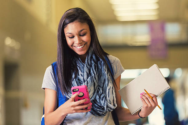 Teenage Hispanic female high school student is smiling as she walks in school hallway. She is looking down at her smart phone and texting someone or using social media. Student has long dark hair, and is wearing a casual printed scarf. She is wearing a backpack and holding a notebook as she walks to class.